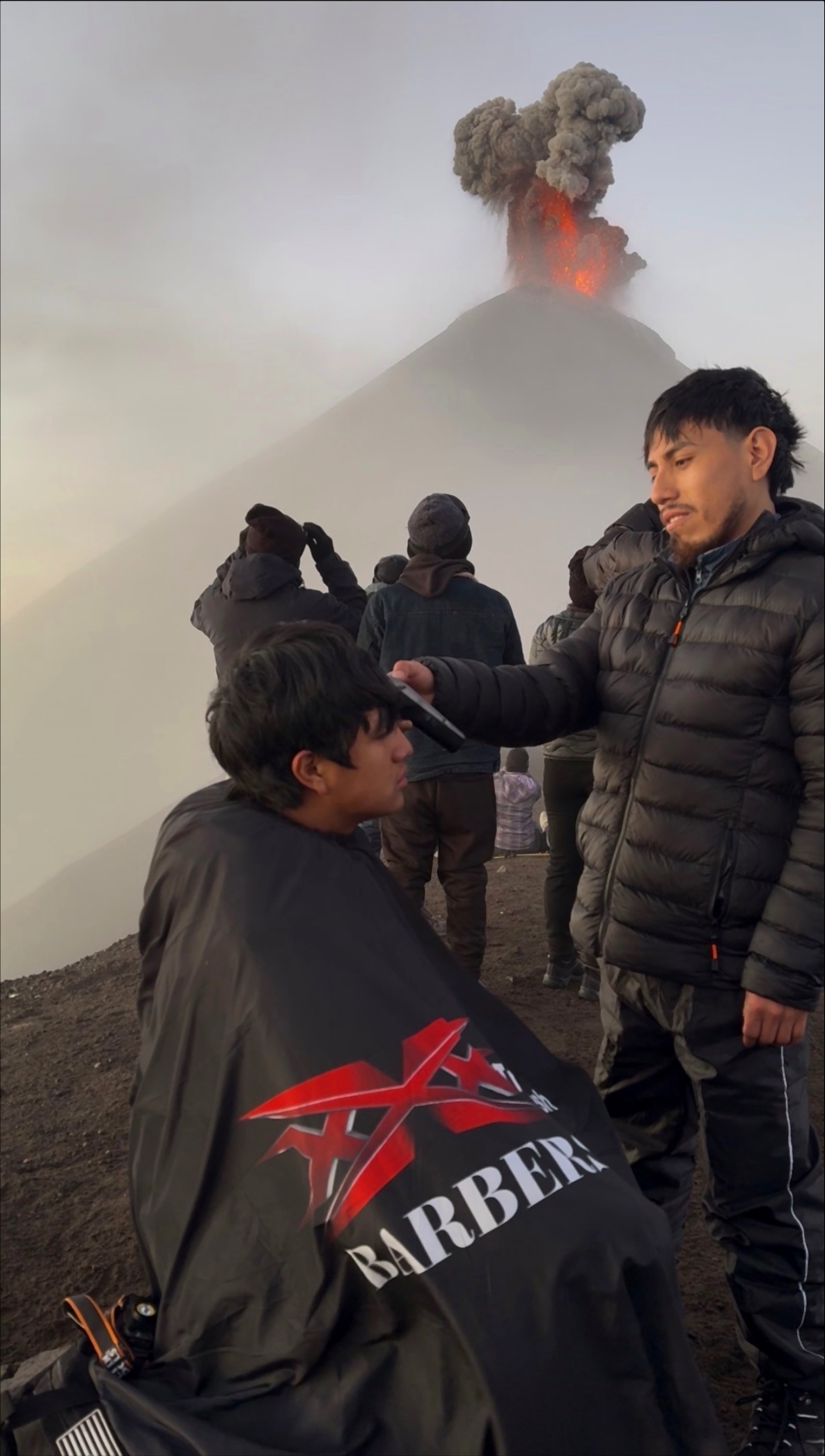 Tavo — Master Barber — cutting hair in front of erupting volcano — full picture